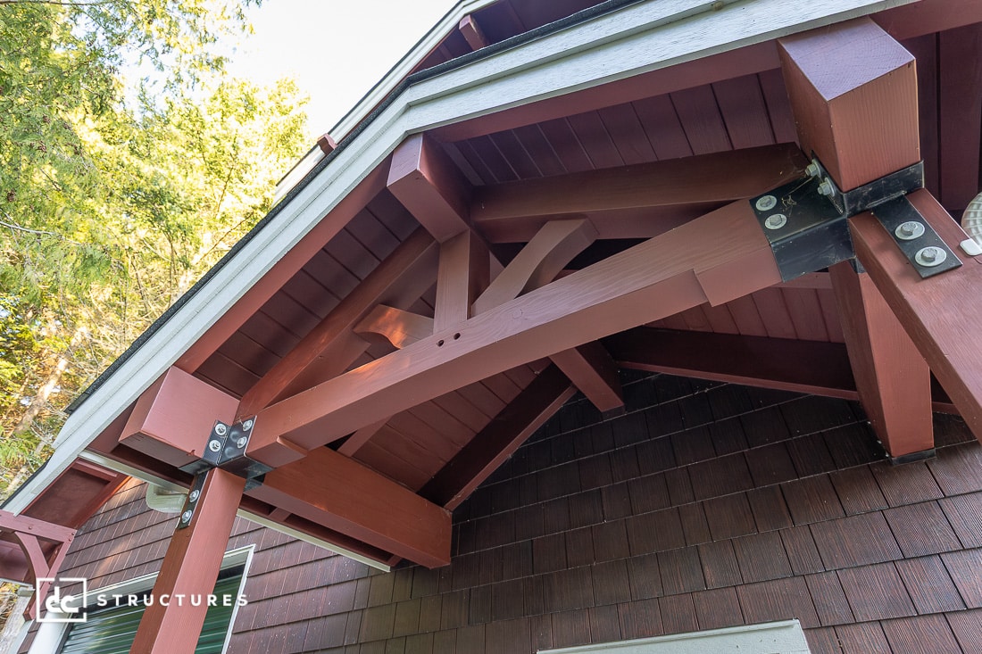 A close-up view of a building's overhanging roof with exposed red wooden beams, metal brackets, and a shingle wall beneath, surrounded by green trees.