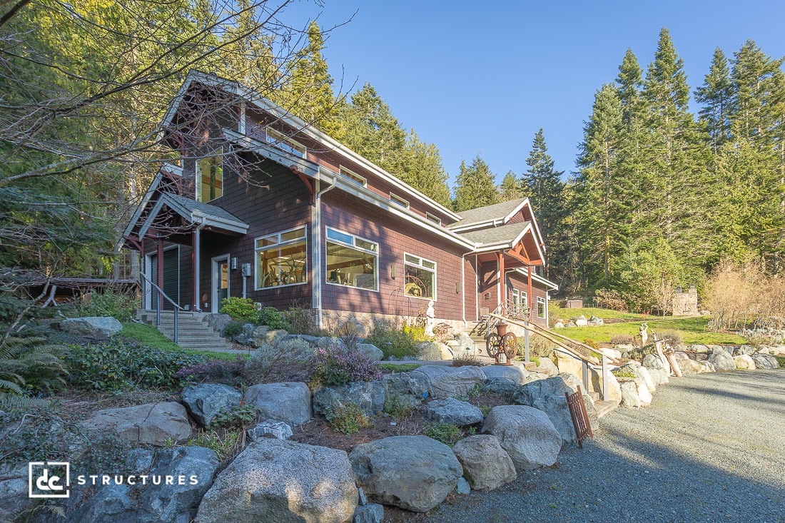 A large wooden house with a steep roof and many windows sits among trees and rocks, with a curved gravel driveway out front.