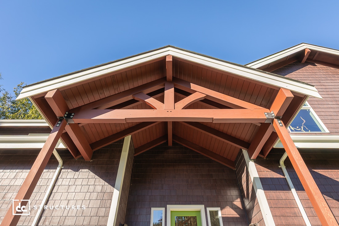 A low-angle view of a house entrance with a peaked wooden porch roof, exposed beams and supports, beneath a clear blue sky.