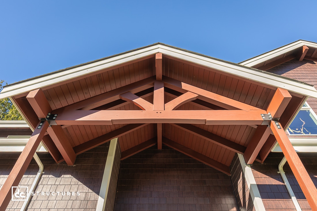 A close-up view of a wooden gabled porch roof with exposed beams and metal brackets, on a brown shingle-sided house, under a blue sky.