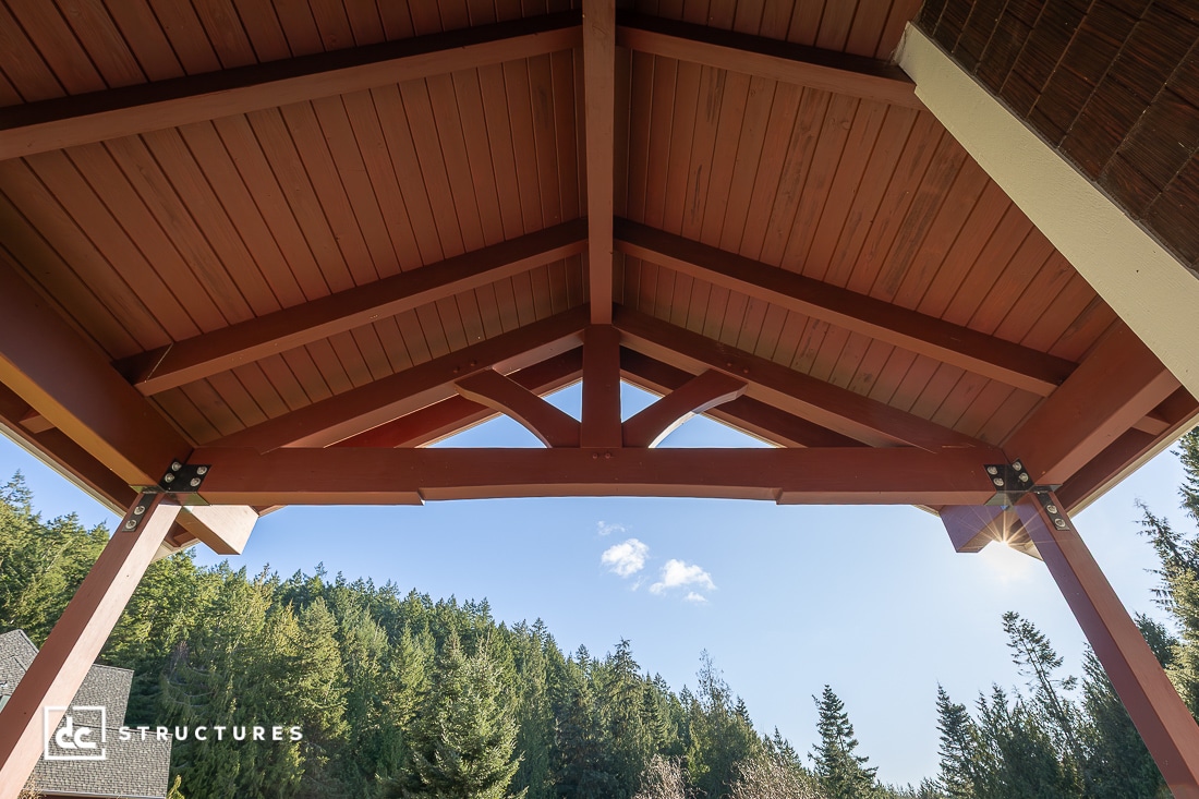 View from beneath a wooden pavilion roof with exposed beams, looking out at a blue sky and forested hillside; metal brackets secure joints.