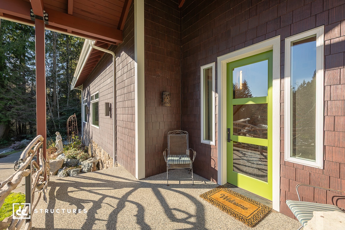 A modern house entrance with brown siding, a green front door with glass panels, a welcome mat, and a metal chair on the porch.