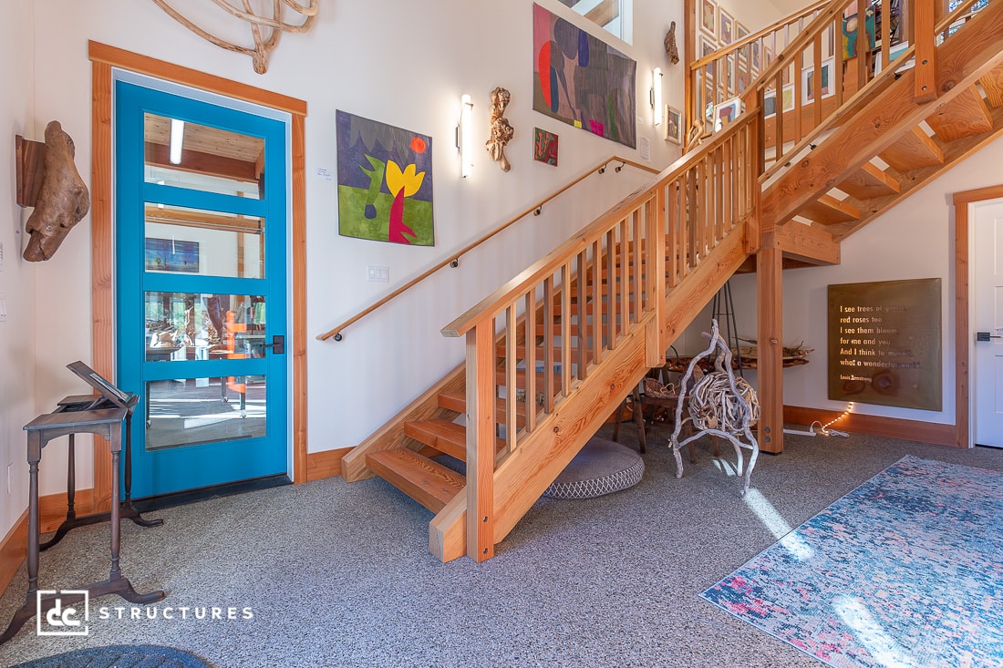 A bright entryway with a blue door, wooden staircase, artwork on the walls, decorative rug, and natural wood accents in sunlight.