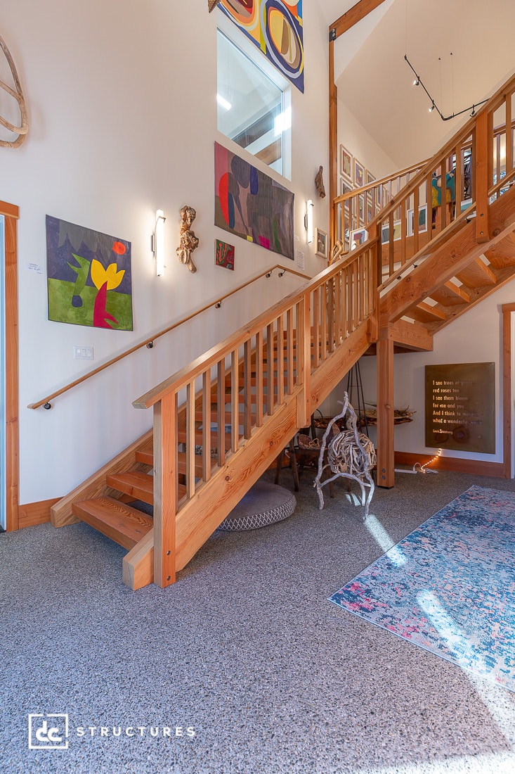 A wooden staircase with railings in a brightly lit entryway, colorful abstract art on white walls, patterned rug, natural light above.