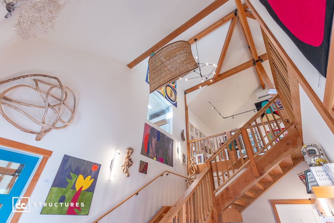 A wooden staircase with railings ascends through a bright, art-filled space with colorful paintings, wood beams, and modern decor.