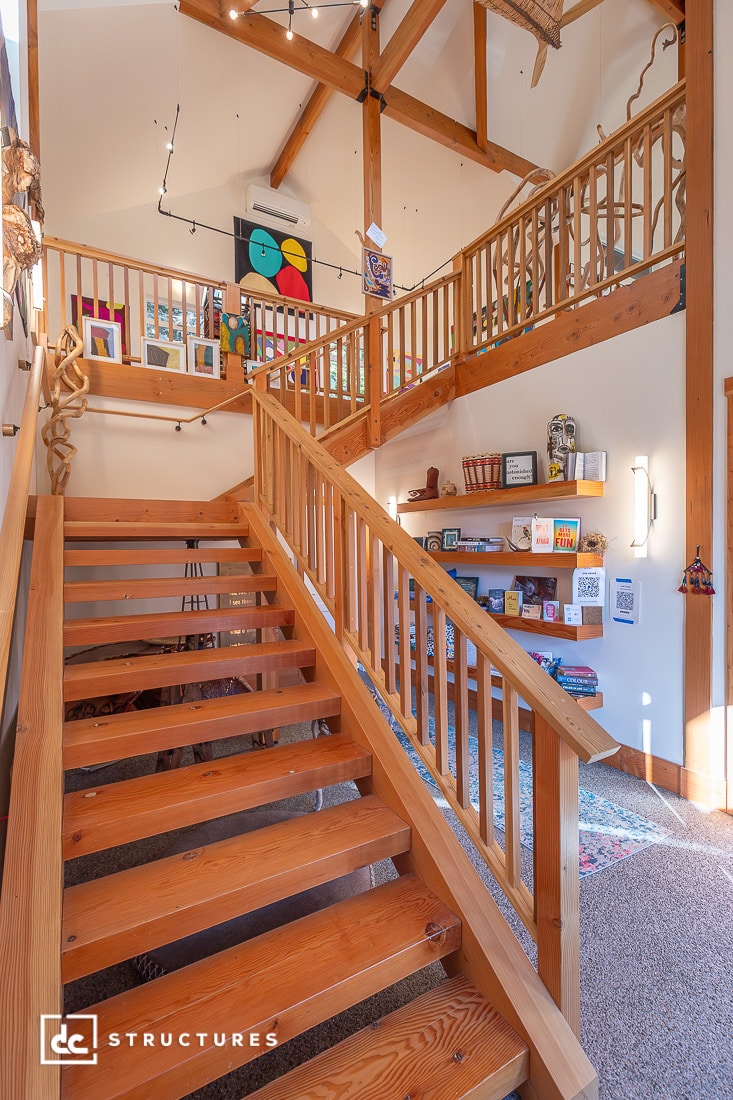 Sunlit wooden staircase with open steps and railings leads to a loft. Art, bookshelves, and decor line walls in a bright space.
