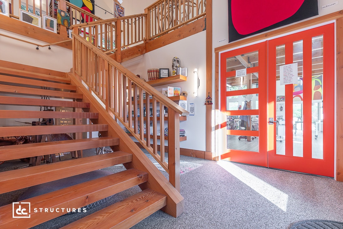 A bright entryway with orange double doors, wooden staircase and railing, wall shelves, large sunny windows, and colorful art.