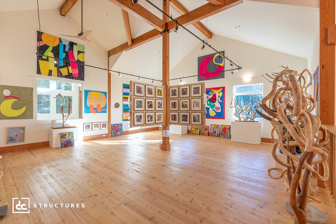 A spacious, sunlit art gallery with wooden floors displays colorful abstract paintings and wooden sculptures on white walls and easels.