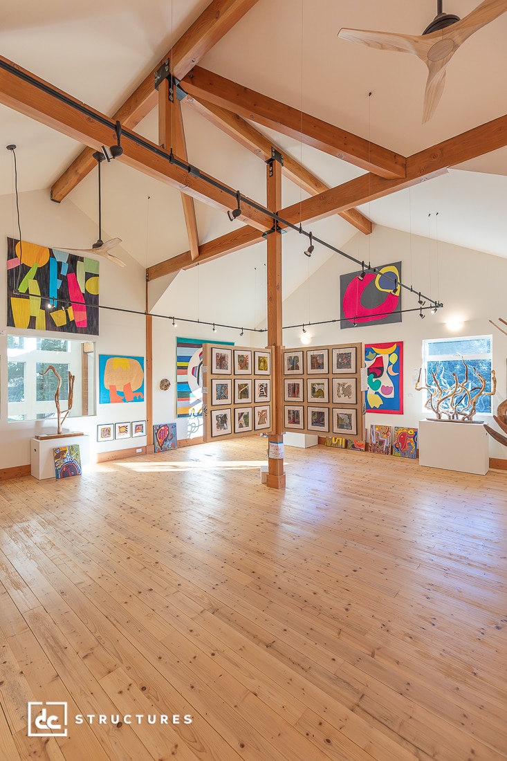 Spacious art gallery with high wooden beams, featuring colorful abstract paintings, framed art, and sculptural pieces on a light wood floor.