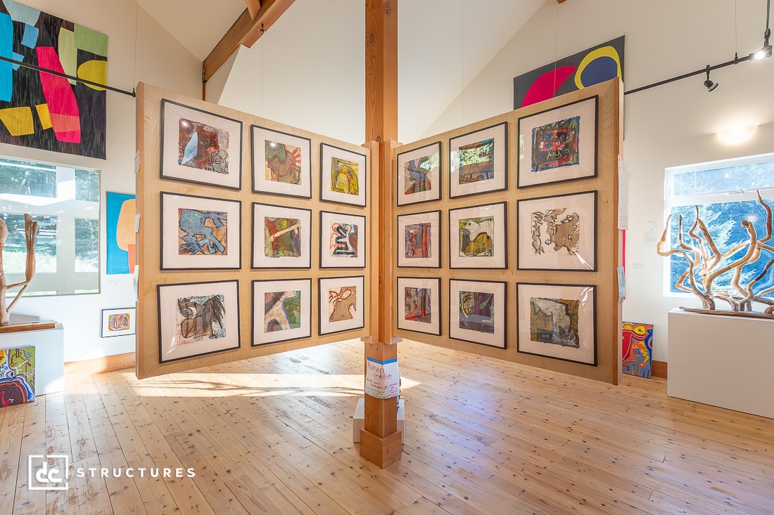 A bright art gallery with wooden floors and high ceilings displays colorful framed artwork on double-sided panels and wooden sculptures.