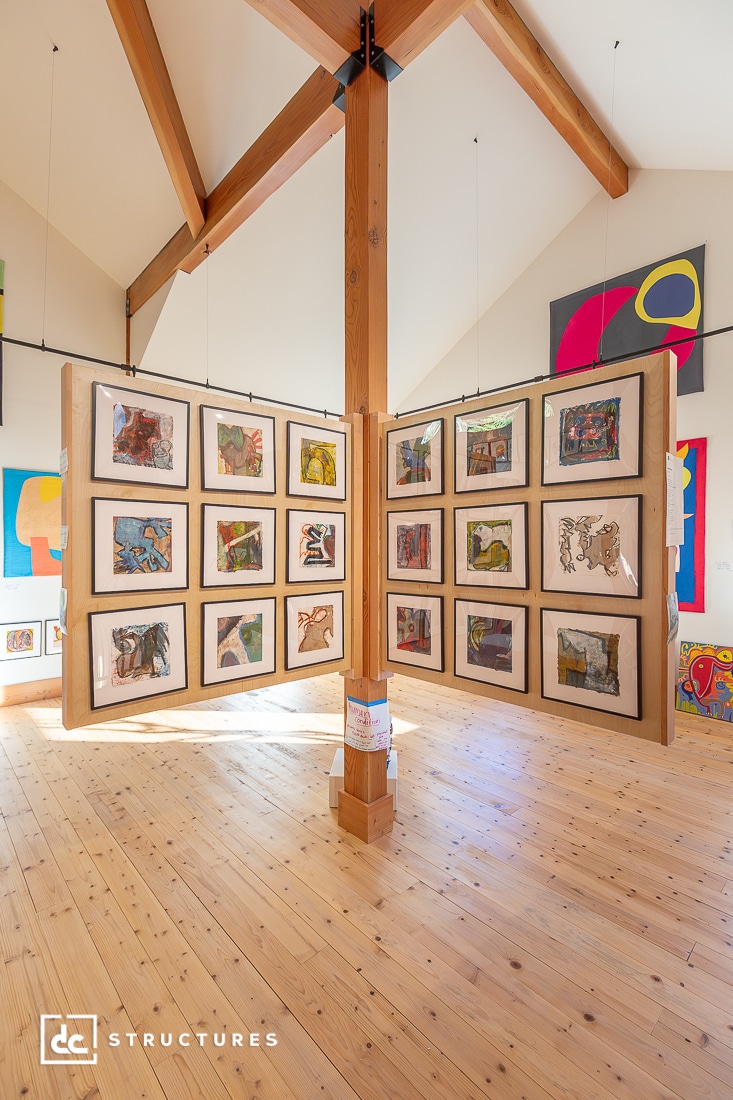 A bright art gallery with wood beams and floor displays framed abstract artworks arranged in two panels forming a corner. Colorful paintings hang on the walls in the background.