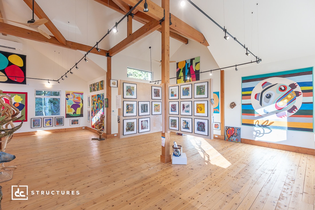 A spacious, well-lit art gallery with wooden floors and beams displays colorful abstract paintings and sculptures on white walls. Large windows let in natural light, highlighting the vibrant artwork.