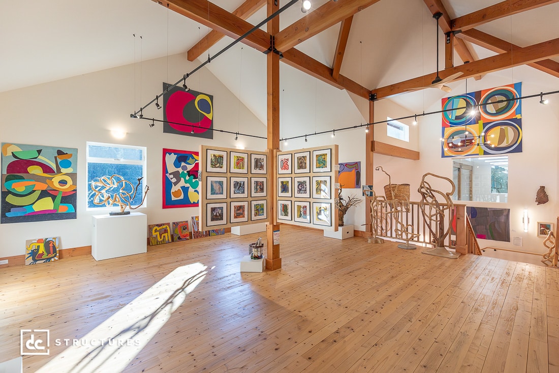 A spacious art gallery with high wooden beam ceilings, large windows, track lighting, and colorful abstract art. Natural light fills the room.