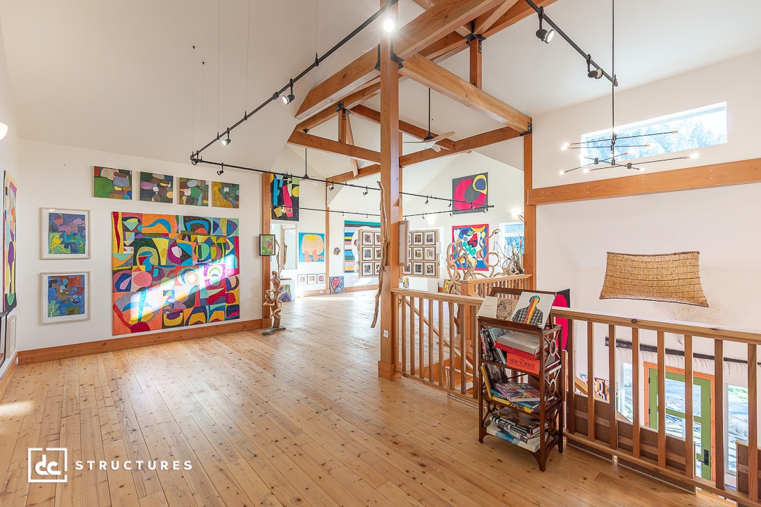 A spacious, bright art studio with wooden floors, exposed beams, and white walls decorated with colorful abstract paintings.