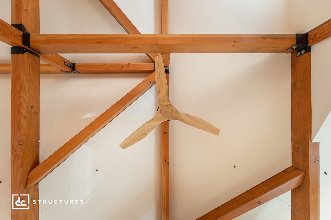 A wooden ceiling fan with three blades is mounted on a timber-framed ceiling with exposed wooden beams and metal joinery.