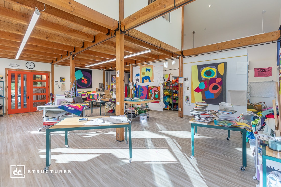 Bright, open art classroom featuring wooden beams, several tables, colorful abstract artwork, shelves with supplies, and large windows.