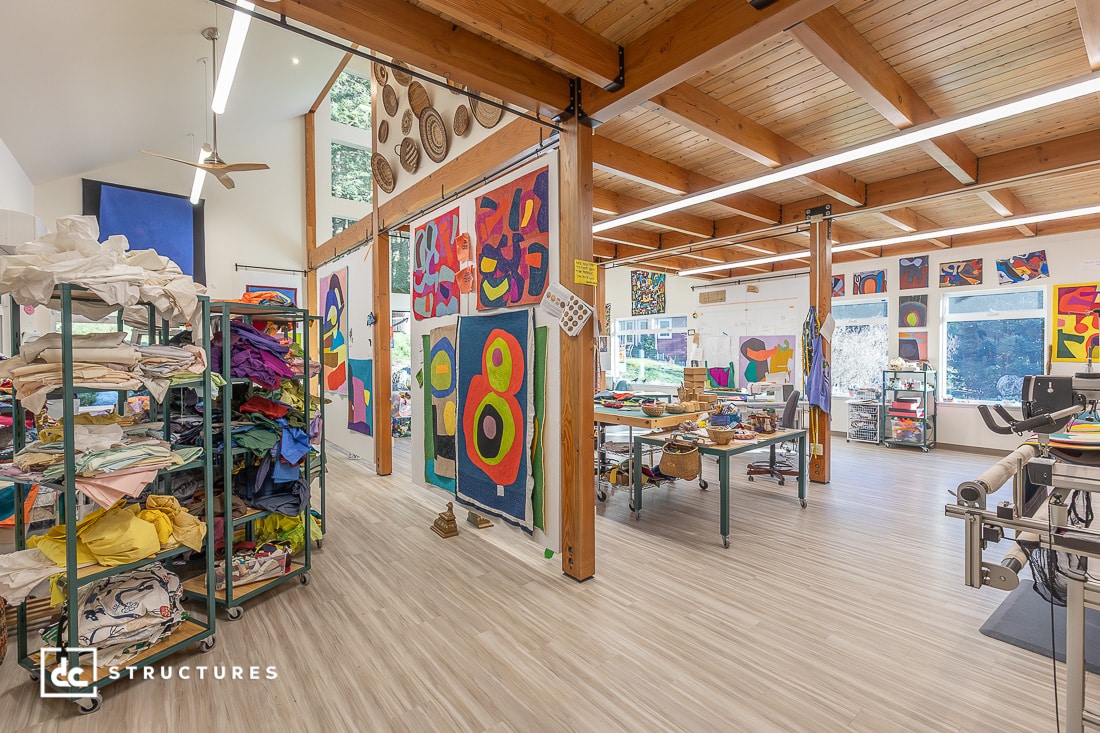 A bright, spacious art studio with wooden beams, abstract paintings, shelves of folded fabrics, worktables, and large windows.