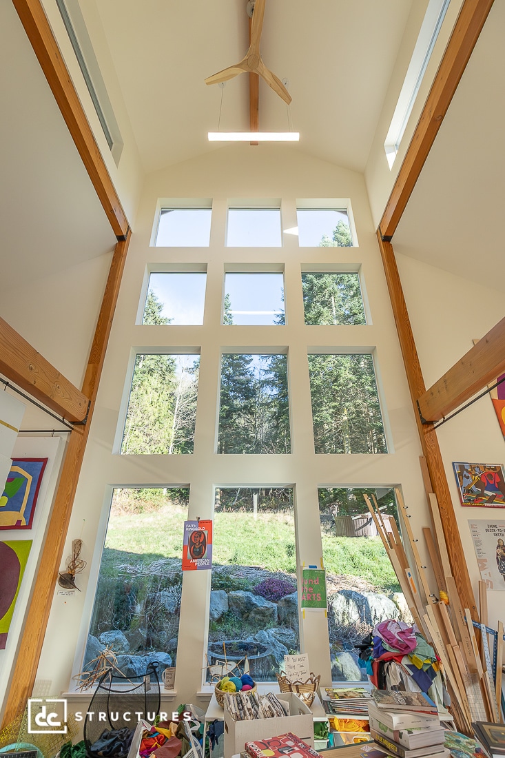 A bright room with tall ceilings, large grid windows, wooden beams, art supplies, and colorful artwork on the walls, overlooking trees.