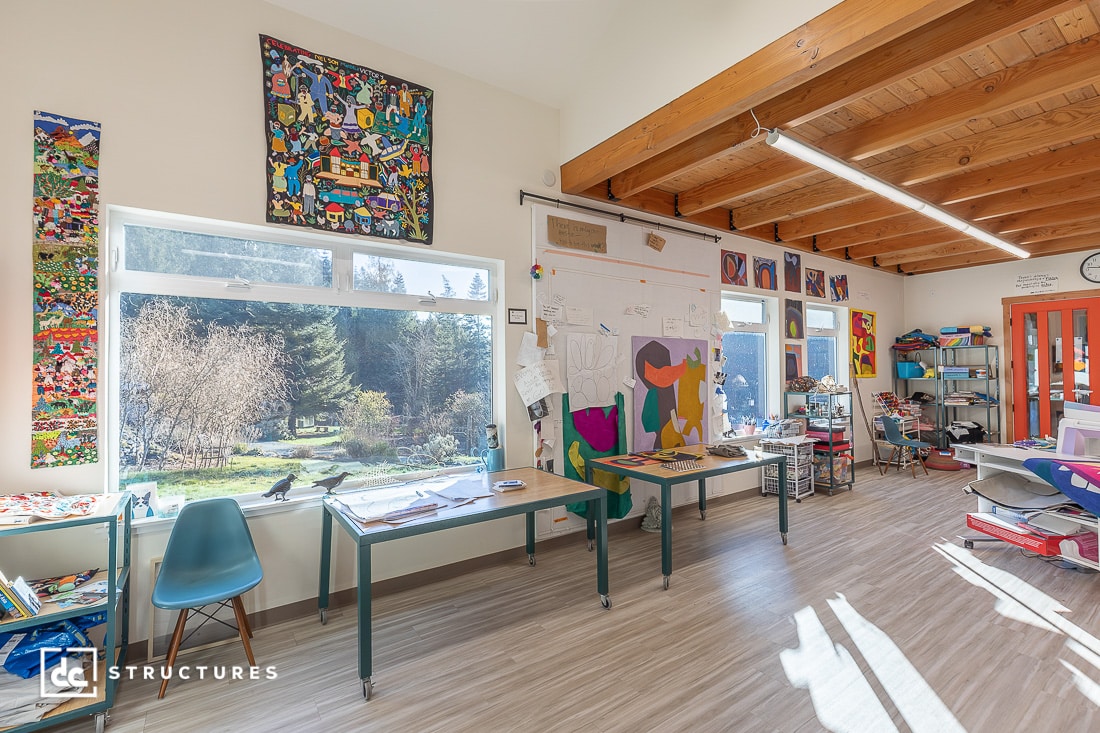 A bright classroom with large windows, colorful artwork on the walls, exposed wooden beams, and desks with art supplies. Trees and greenery are visible outside. The space appears creative and inviting.