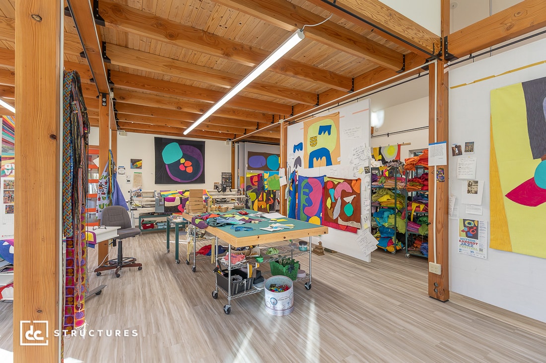 A bright, open art studio with wooden beams, colorful abstract quilts on the walls, a large worktable, and shelves of supplies.