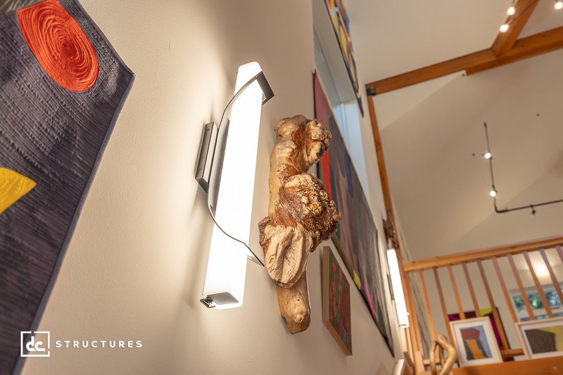 A modern wall sconce with a wooden accent on a wall decorated with colorful art; wooden railing and framed artwork in the background.