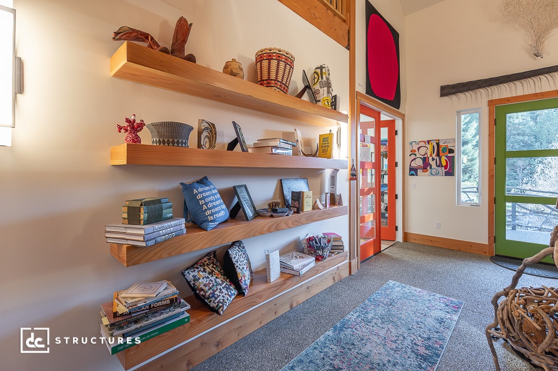 A cozy room with three wooden floating shelves holding books, baskets, and decor. There's a patterned rug, framed art, and a green door with large windows letting in natural light.