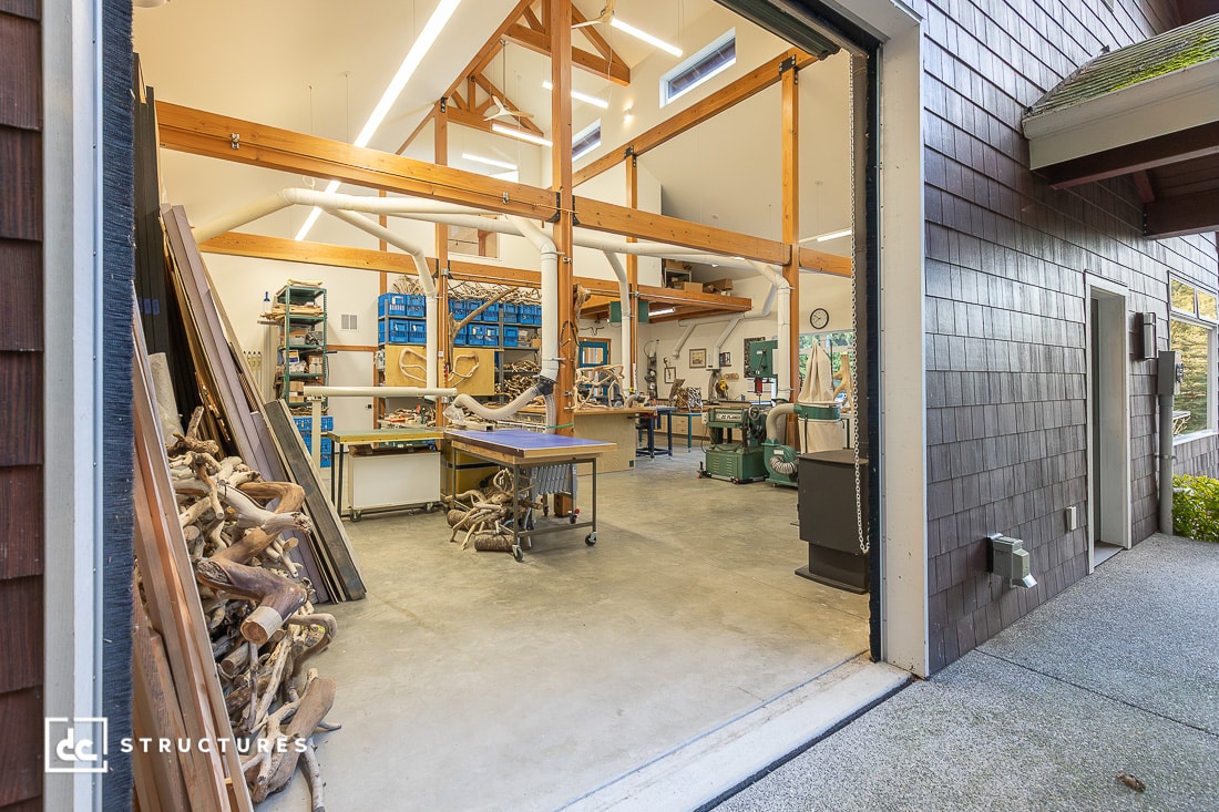 A spacious woodworking shop with a large open garage door, featuring woodworking tools, lumber, and natural light.
