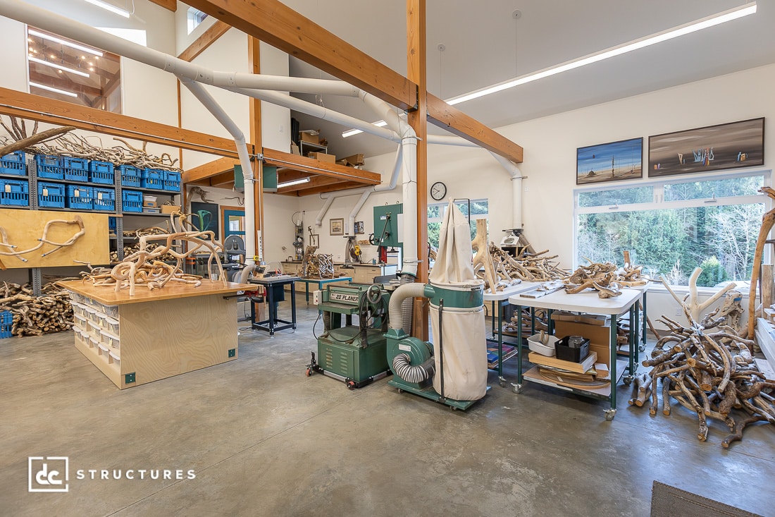 A spacious wood workshop with exposed beams, large machinery, worktables, blue bins, and driftwood piles. Sunlight streams in.