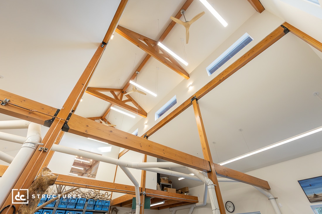 Interior view of a modern building with exposed wooden beams, high ceilings, large windows, and contemporary lighting fixtures. The structure features wood and metal elements.