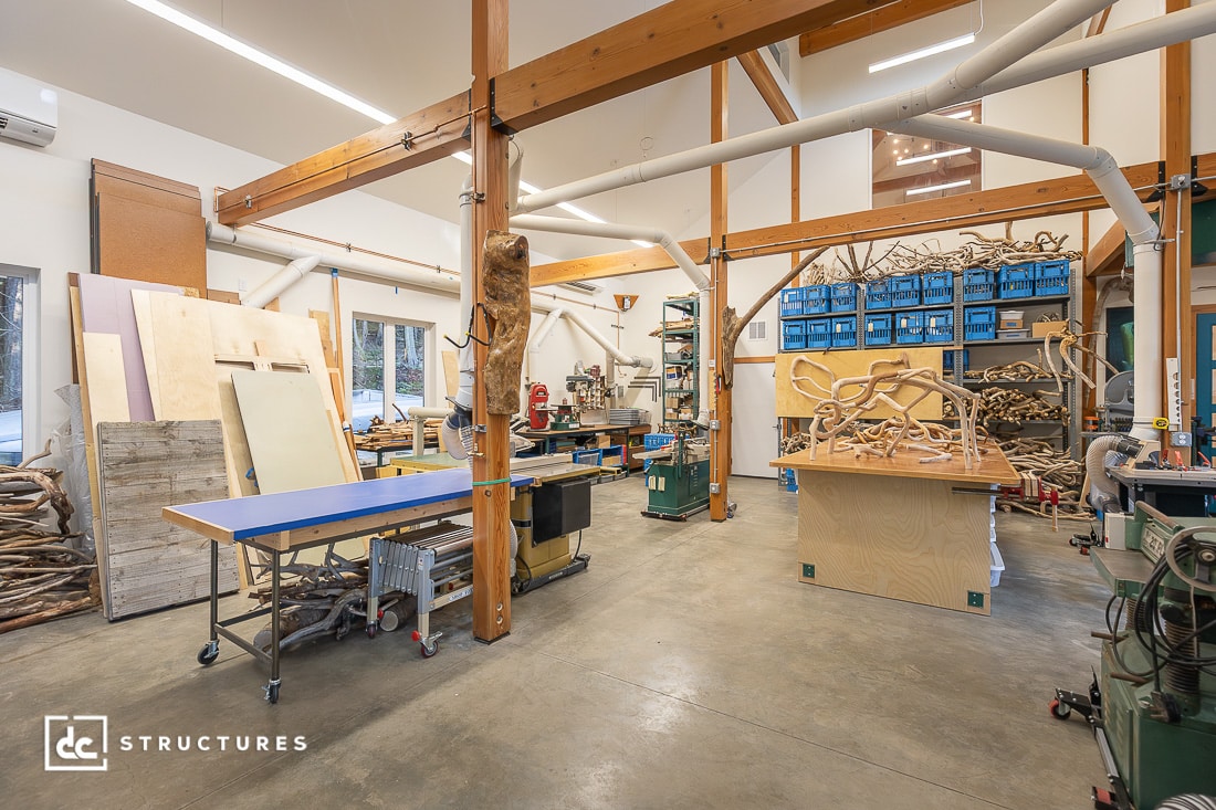 A spacious woodworking workshop with high ceilings, large windows, work tables, woodworking machines, and organized shelves.