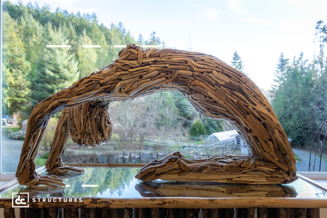 A sculpture of intertwined driftwood forms a human in a backbend yoga pose, displayed indoors by a window with forest views outside.