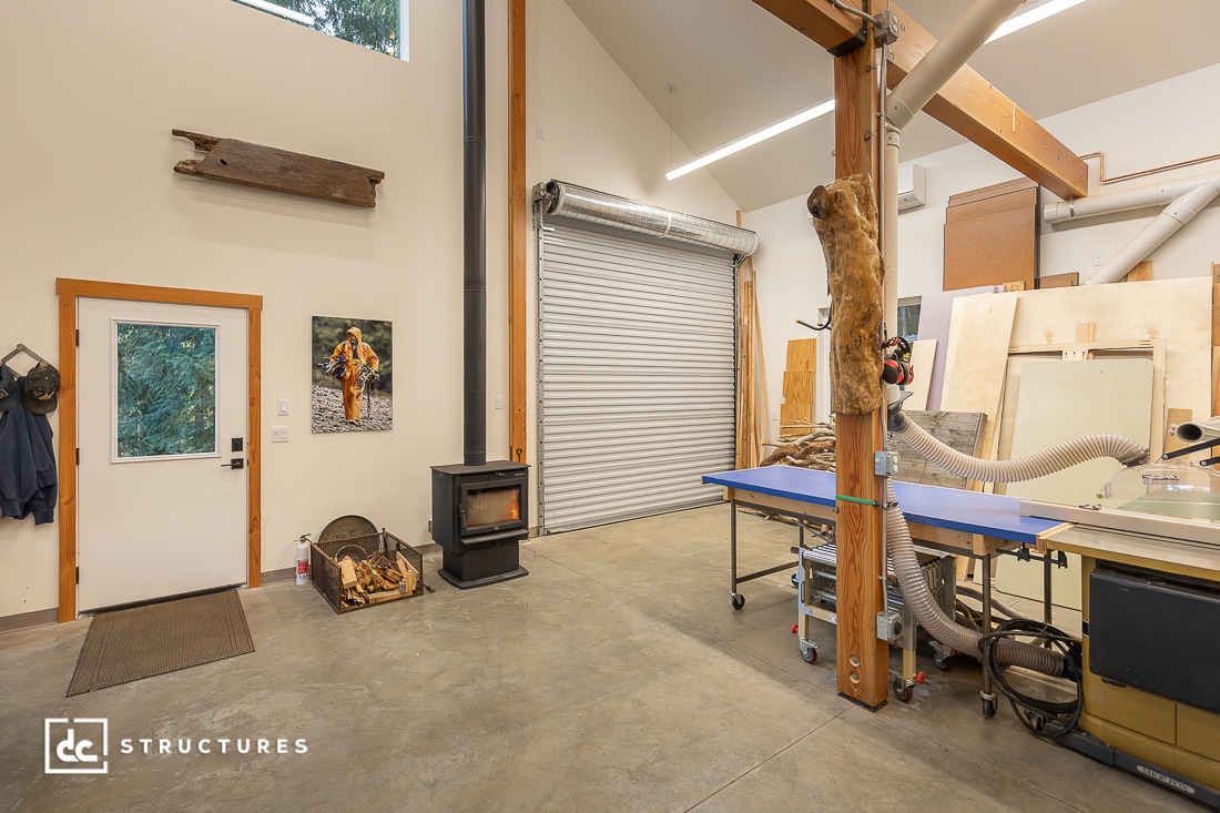 A spacious woodworking workshop with a roll-up garage door, wood stove, firewood, workbenches, tools, beams, and large windows.