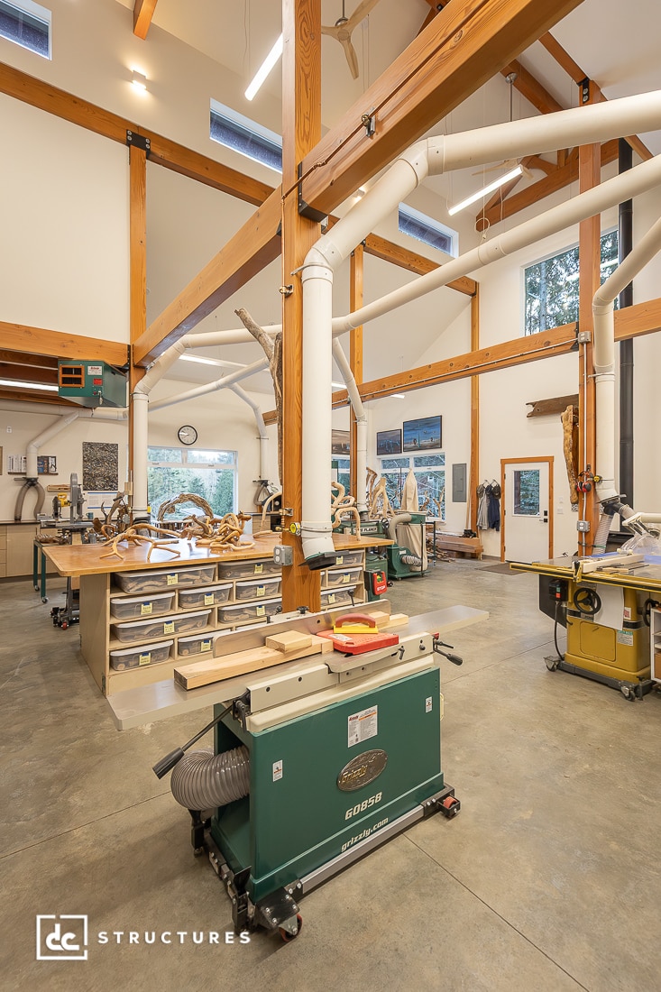 A spacious, well-lit woodworking shop with high ceilings, exposed beams, large windows, worktables, machinery, and organized tools.