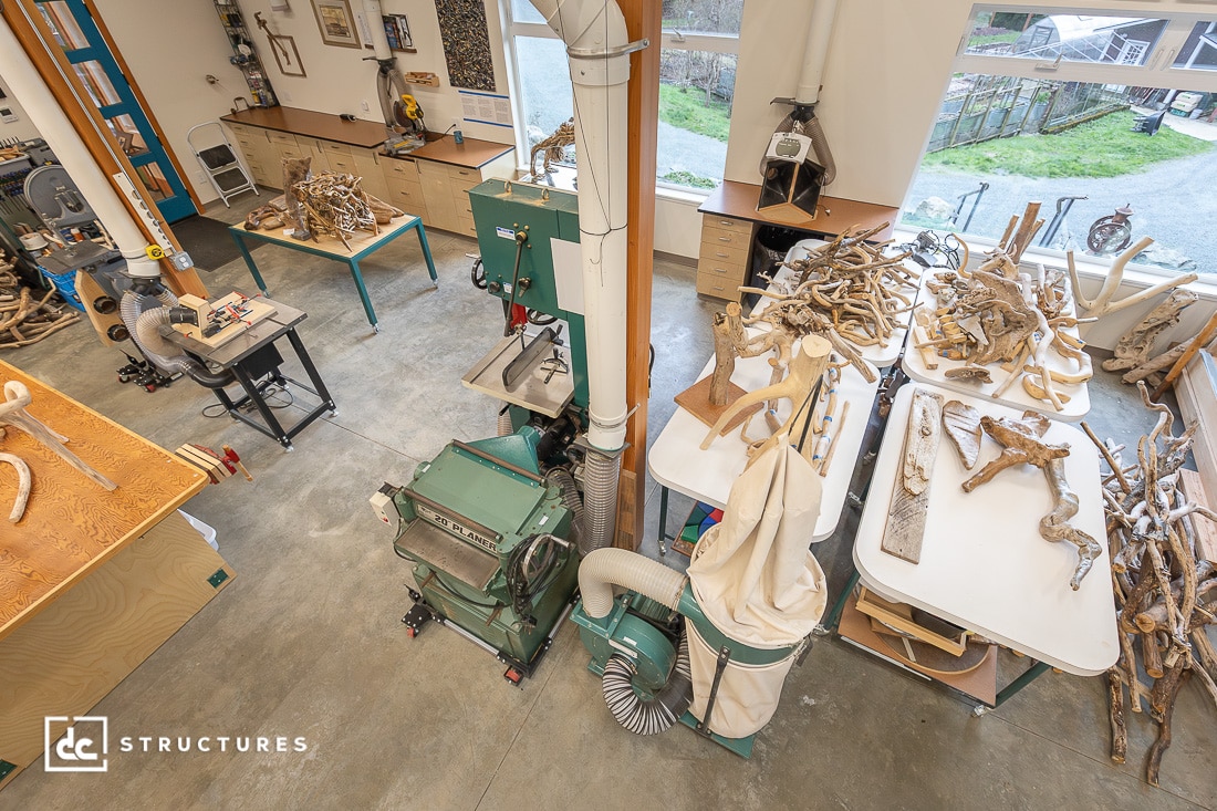 A bright, organized woodshop with woodworking machines, driftwood sculptures on tables, and large windows overlooking a grassy yard.