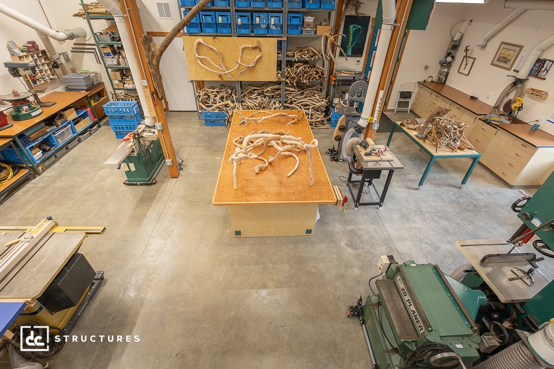A woodworking shop with a central workbench covered in twisted wooden branches, surrounded by machines, tools, and storage bins.