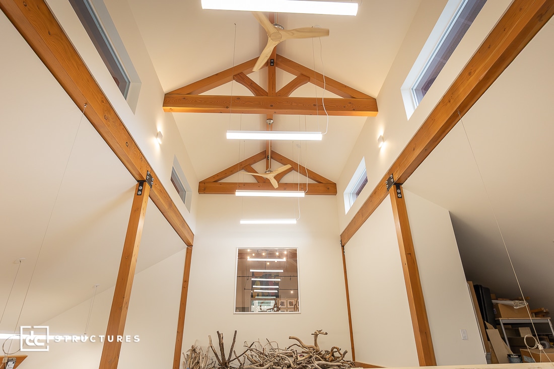 Interior view of a modern building with exposed wooden beams, vaulted ceiling, large windows, hanging lights, and driftwood display.