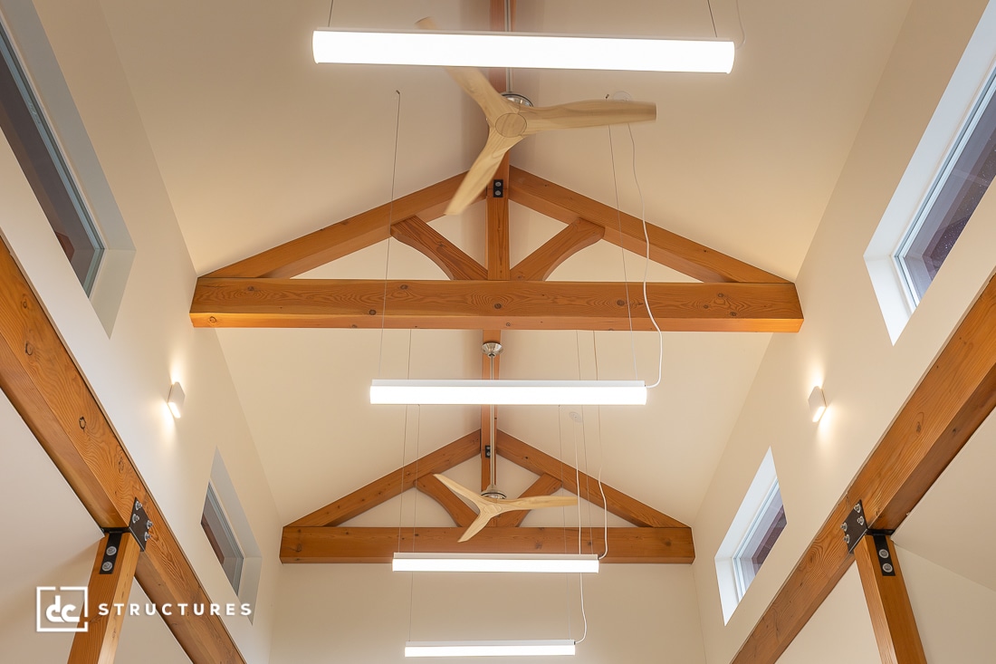 Exposed wooden beams and trusses on a vaulted ceiling with modern light fixtures and a wooden ceiling fan. The lighting creates a bright and airy atmosphere.