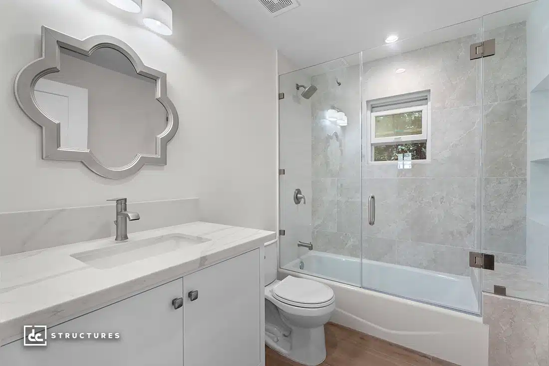 Modern bathroom with a white vanity, marble countertop, unique mirror, toilet, bathtub with glass shower, wood floor, small window.