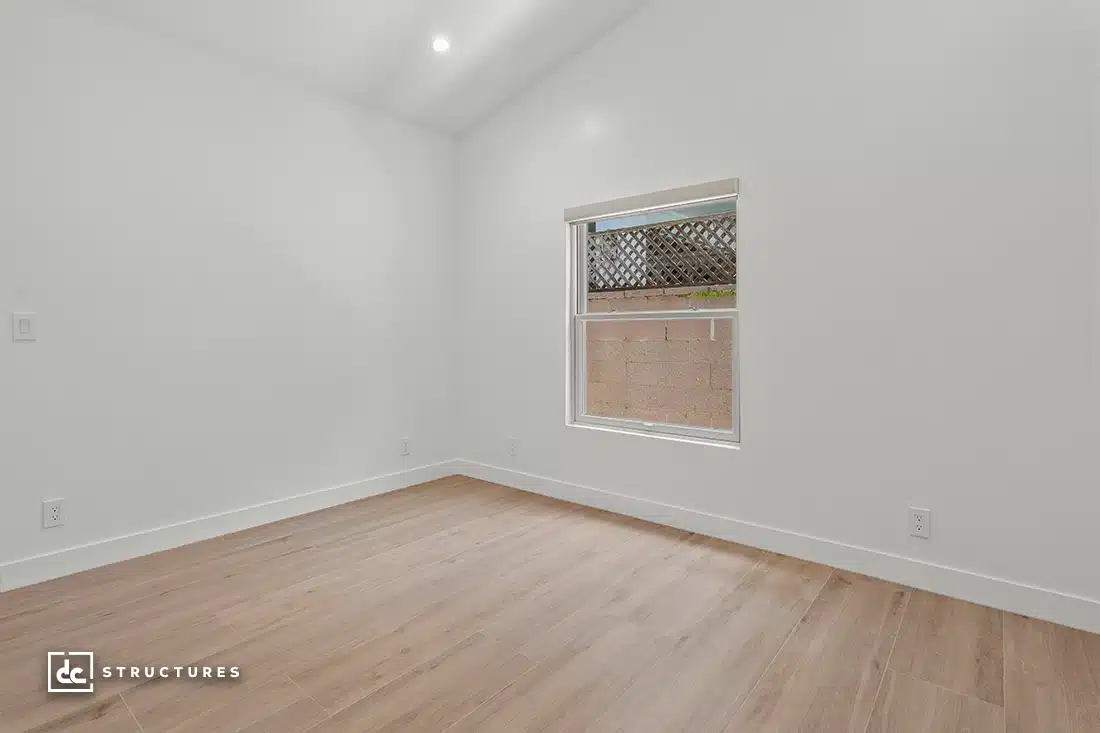 A minimalist empty room with white walls, a light wood floor, a single window showing part of a brick wall and wooden lattice, and recessed ceiling lights.