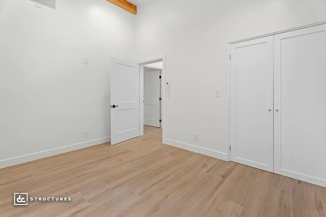 Minimalist, empty room with light wood flooring, white walls, double closet with white doors, open door to hallway, wooden ceiling beam.
