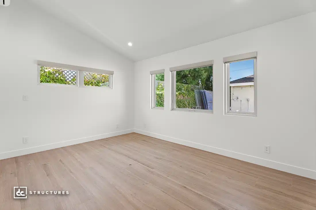 A bright, empty room with white walls, light wood floor, four windows showing greenery, and a sloped ceiling with recessed lighting.