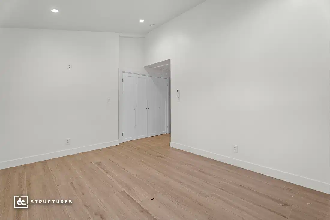 A bright, empty room with white walls, wood flooring, a small closet with double white doors, and recessed lighting on the ceiling.