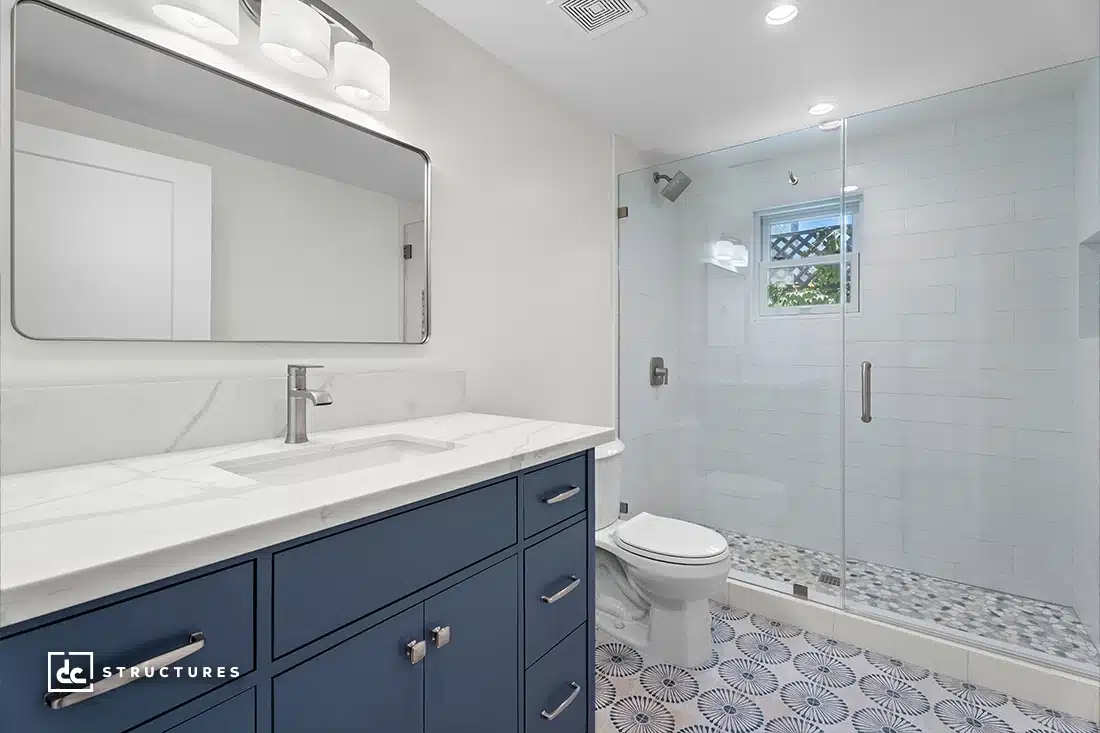 Modern bathroom with a blue vanity, white countertop, large mirror, overhead lights, glass shower, patterned tile, and window.