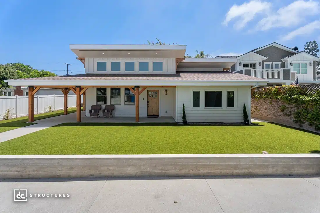 Modern two-story house with flat roof, large windows, wooden front porch, well-manicured lawn, and two chairs on the porch.