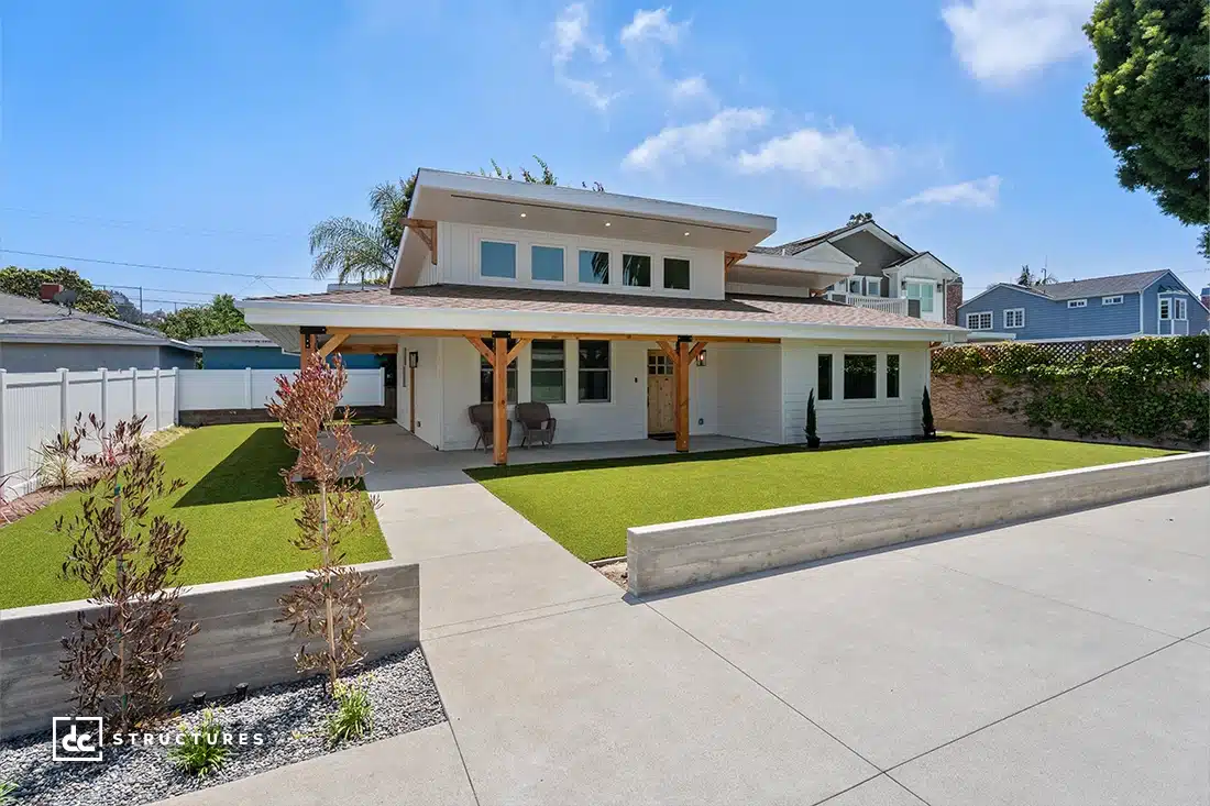 Modern two-story house with a wide driveway, covered front porch, minimalist landscaping, and artificial turf lawn under blue sky.
