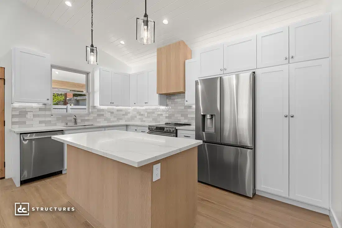 Modern kitchen with white cabinets, stainless steel appliances, light wood island with white countertop, pendant lights, and window.