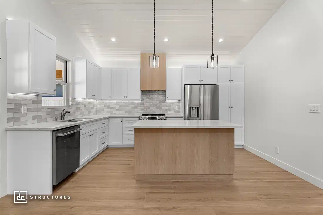 Modern kitchen with white cabinets, stainless steel appliances, light wood island with white countertop, tile backsplash, wood flooring, and pendant lights.
