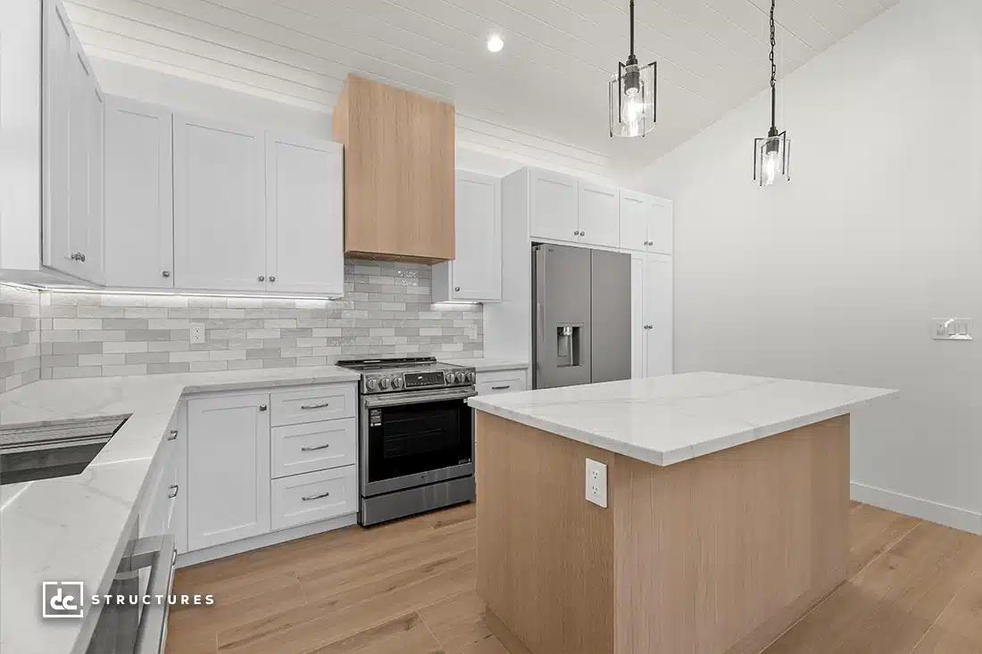 Modern kitchen with white cabinets, light wood accents, stainless steel appliances, marble countertops, island, and wood flooring.