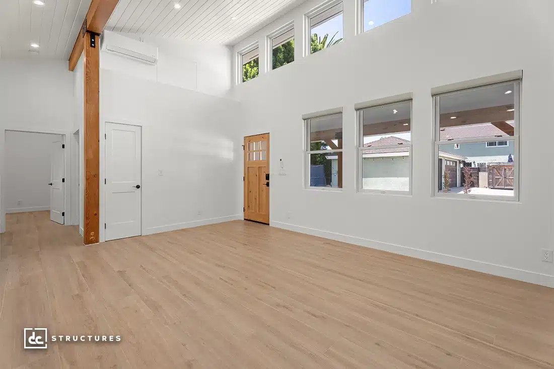 A bright, modern empty room with light wood floors, white walls, large windows, a wooden front door, and exposed beams.