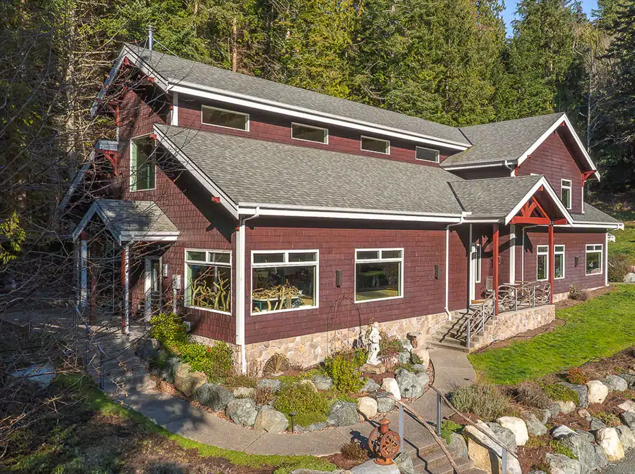 A large two-story house with dark red siding and a gray roof, stone foundation, large windows, surrounded by rocks and trees.